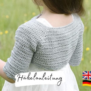 Puede incluir: Bolero de ganchillo gris claro con mangas tres cuartos. El bolero se lleva sobre un vestido blanco. La imagen incluye el texto "Häkelanleitung". El fondo es un campo verde borroso con flores amarillas.