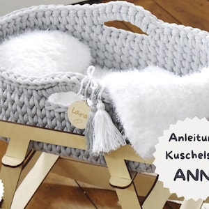 May include: A gray, chunky-knit baby bassinet with a white fluffy interior and accents. The bassinet sits on a light wood rocking base. A wooden tag with the name "Lara" hangs from the side. The text "Anleitung Kuschelset ANNI" is visible.