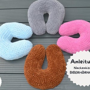 Puede incluir: Cinco almohadas de cuello de ganchillo en forma de U en azul claro, gris, rosa y marrón. Las almohadas están dispuestas sobre una superficie gris. La imagen también incluye una bandera alemana y texto en alemán.