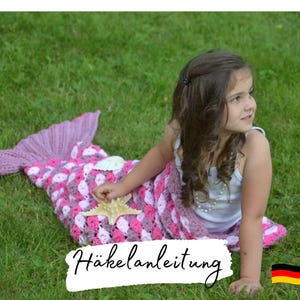 May include: A young girl wearing a white tank top sits on a green grassy lawn. She is wrapped in a pink and purple crocheted mermaid tail blanket. The blanket has a shell and starfish embellishment. The text "Häkelanleitung" is visible in the image.