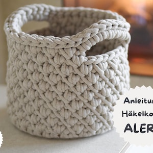 May include: A beige, hand-crocheted basket with two handles. The basket has a textured, woven appearance and a round shape. The text "Anleitung Häkelkorb ALERA" is visible on a white label. The basket is likely for storage or decoration.