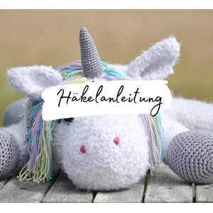 May include: A white crocheted unicorn with a rainbow mane and gray hooves. The unicorn is lying down on a wooden surface. The text "Häkelanleitung" is written on a white background.