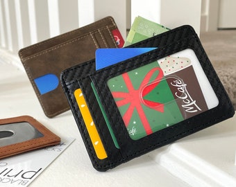 May include: Two black leather wallets with a carbon fiber pattern. One wallet has a clear window displaying a gift card with a red bow and the text "McCafe". The other wallet has a blue card inside.