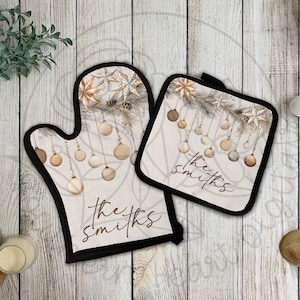 May include: A matching oven mitt and pot holder set with a festive design. The beige fabric features hanging ornaments, stars, and the text "the smiths" in a script font. The set has a black trim.