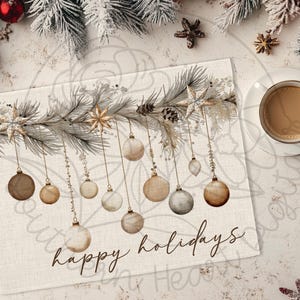 May include: A rectangular placemat with a watercolor design featuring hanging ornaments and the text "happy holidays." The ornaments are in shades of brown and gold, suspended from a branch with stars and pinecones. A cup of coffee sits on a white saucer.