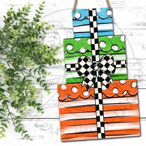 May include: A decorative wooden sign featuring three stacked gift boxes in blue, green, and orange. Each box has a unique pattern, including stripes and polka dots. A black and white checkered bow and ribbon accent the gifts.