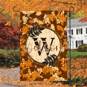May include: A fall-themed garden flag with a brown circle containing the letter "W" and the word "Watson" in a white font. The circle is surrounded by orange, yellow, and brown leaves.