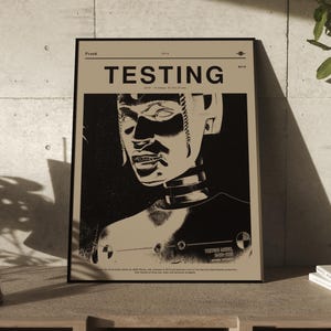 Puede incluir: Un póster enmarcado con la palabra "TESTING" en letras mayúsculas grandes. El póster presenta una imagen en blanco y negro de la cabeza y la parte superior del cuerpo de un maniquí de prueba de choque. El póster se muestra sobre una superficie de madera.