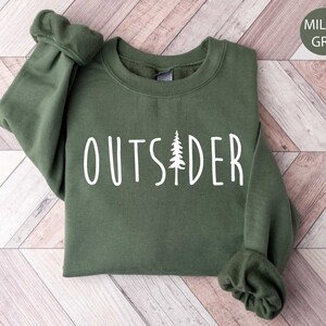 May include: Military green sweatshirt with the word "OUTSIDER" in white, featuring a tree graphic. The sweatshirt has a crew neck and long sleeves with ribbed cuffs. The background is a light wood pattern.