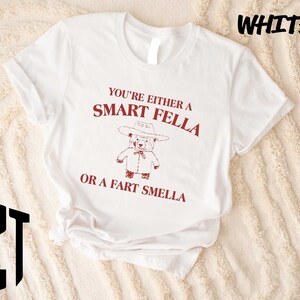 May include: A white short-sleeved t-shirt featuring a red graphic of a bear wearing a cowboy hat. The text above the bear reads YOU'RE EITHER A SMART FELLA and the text below the bear reads OR A FART SMELLA. The word WHITE is printed in the top right corner.