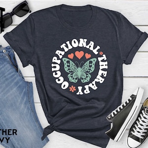 May include: A navy blue t-shirt with the text "Occupational Therapy" in a white circle with a butterfly and hearts. The shirt is on a wooden surface with a pair of jeans and a pair of black sneakers.