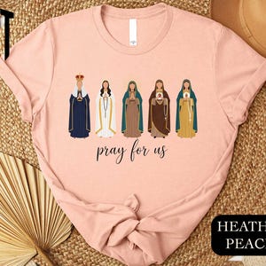 A pink t-shirt with a graphic design featuring five stylized female figures in different religious attire, along with the text " Prayer for us" printed on the shirt.