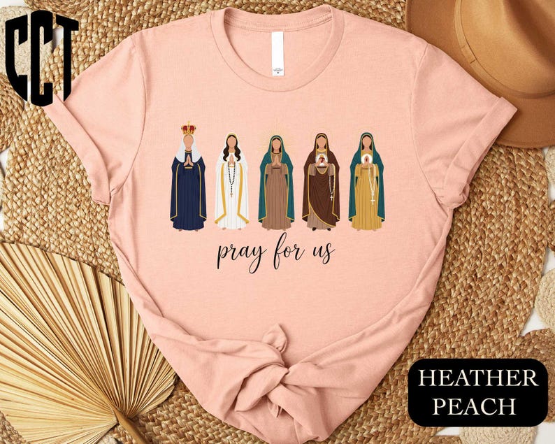 A pink t-shirt with a graphic design featuring five stylized female figures in different religious attire, along with the text " Prayer for us" printed on the shirt.