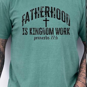 May include: A teal green t-shirt with the text "FATHERHOOD IS KINGDOM WORK" and a cross graphic. The text "proverbs 22:6" is below the main text. The shirt is short-sleeved.
