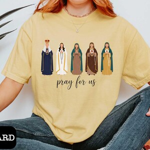 A person wearing a yellow t-shirt with a graphic design of five religious figures and the text "Pray for us" printed on it.