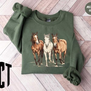 May include: A green sweatshirt with a graphic of three horses. The horses are brown, white, and brown. The sweatshirt has a crew neck and long sleeves.