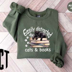 May include: A green sweatshirt with the text "Easily distracted by cats & books" and a black cat sleeping on a stack of books.