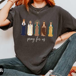 A person wearing a gray t-shirt with a graphic design of five religious figures and the text "Pray for us" printed on it.