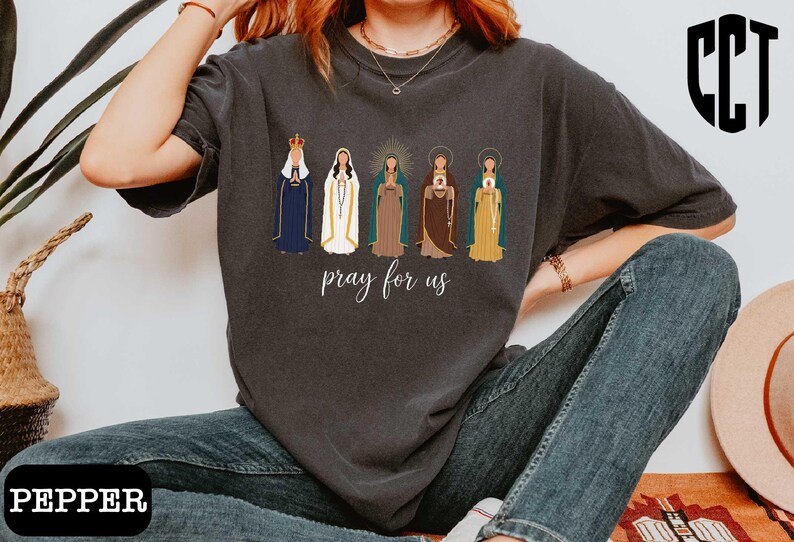 A person wearing a gray t-shirt with a graphic design of five religious figures and the text "Pray for us" printed on it.
