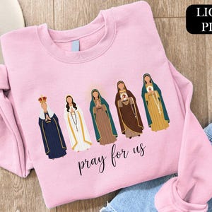 A pink t-shirt with a graphic design of five religious figures, including Mary, the Virgin Mary, and other saints, along with the text "Pray for us" printed on it.