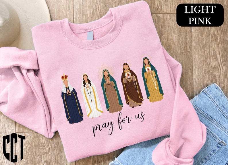 A pink t-shirt with a graphic design of five religious figures, including Mary, the Virgin Mary, and other saints, along with the text "Pray for us" printed on it.