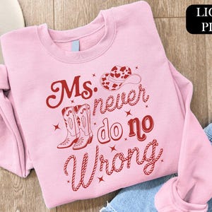 May include: Light pink sweatshirt with the text "Ms. never do no Wrong" in red script. The design includes a cowboy hat and boots. The sweatshirt is folded on a wooden surface.