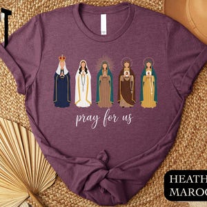 a purple t-shirt with a graphic design of five religious figures, including Mary, the Virgin Mary, and other saints, along with the text " Prayer for us" printed on it.