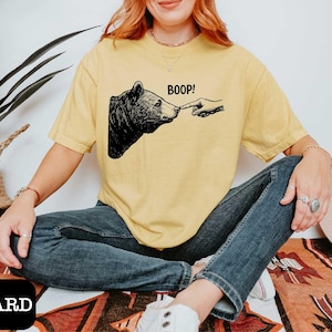 May include: Mustard-colored t-shirt featuring a black and white graphic of a bear being poked on the nose with a finger, with the word "BOOP!" above the bear. The shirt is a relaxed fit.