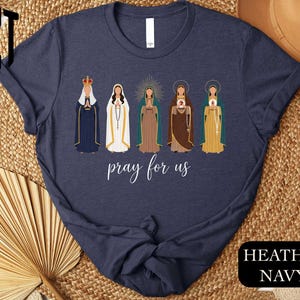 A navy blue t-shirt with a graphic design featuring five different religious figures, including a robed figure, a figure with a halo, and other figures in various colors and poses. The text "pray for us" is displayed below the figures.