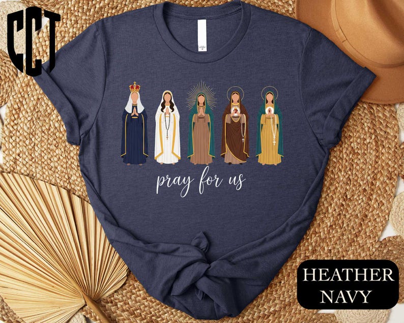 A navy blue t-shirt with a graphic design featuring five different religious figures, including a robed figure, a figure with a halo, and other figures in various colors and poses. The text "pray for us" is displayed below the figures.