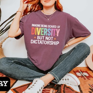 May include: Berry-colored t-shirt with the text "IMAGINE BEING SCARED OF DIVERSITY BUT NOT DICTATORSHIP" in rainbow colors. The shirt is worn with blue jeans and white sneakers. A straw hat and a woven basket are in the background.