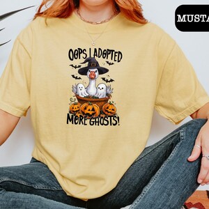 May include: A mustard yellow t-shirt featuring a graphic of a goose wearing a witch hat, sitting in a basket with two ghosts and three jack-o'-lanterns. The text reads "OOPS I ADOPTED MORE GHOSTS!" with small black bats flying around the design.