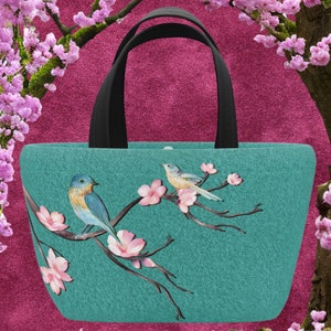 Pode incluir: Uma bolsa tote de feltro turquesa com alças pretas apresenta um design floral com dois pássaros. Os pássaros são um pardal azul e um pássaro amarelo com detalhes pretos. As flores são brancas com detalhes rosa.