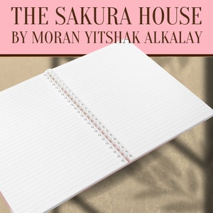 May include: An open spiral bound notebook with white lined pages and a pink cover. The notebook is lying open on a brown surface. The text "THE SAKURA HOUSE BY MORAN YITSHAK ALKALAY" is printed in black on a pink background at the top of the image.