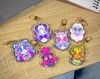 May include: A collection of seven acrylic keychains featuring colorful, cartoon monster girl designs. Each keychain has a gold-colored clasp. The characters have various hair colors, including purple, pink, blue, and green, and are set against a clear background.