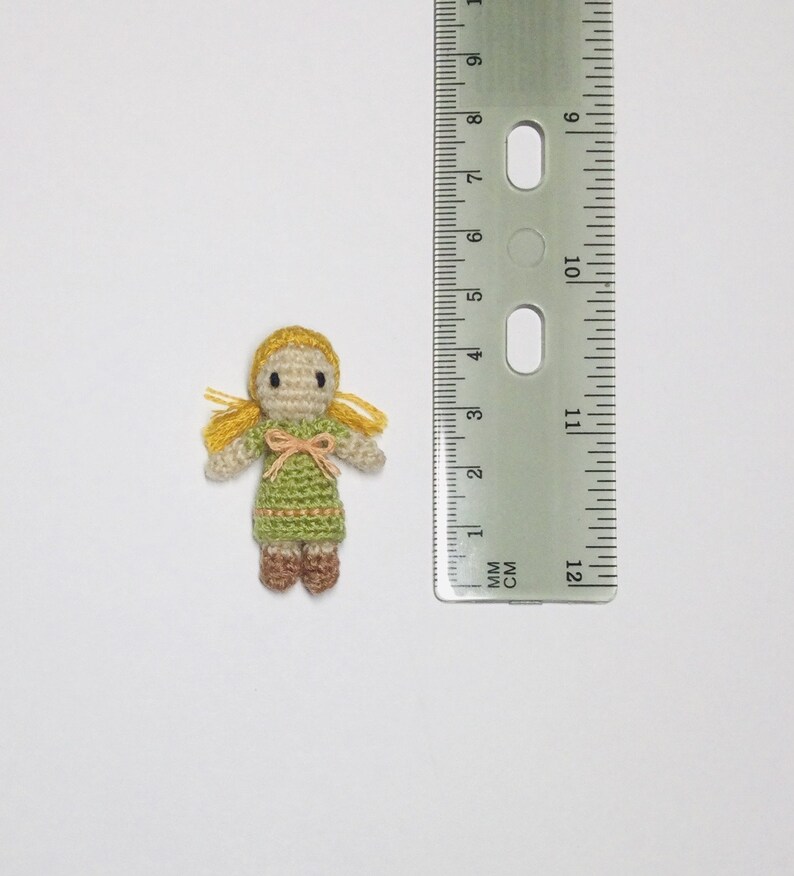 K&ouml;nnte beinhalten: Eine kleine geh&auml;kelte Puppe mit blonden Haaren, einem gr&uuml;nen Kleid und braunen Schuhen. Die Puppe ist etwa 2 cm gro&szlig;.