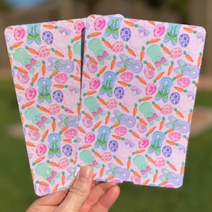 May include: Two light purple Easter-themed notebooks with a repeating pattern of bunnies, carrots, and Easter eggs. The notebooks are held in front of a green grassy background.
