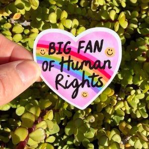 May include: A heart-shaped sticker with the text "BIG FAN OF Human Rights" in black, rainbow, and white. The sticker has a pink background, a rainbow arch, and smiling faces. The sticker is held against a background of green and yellow leaves.
