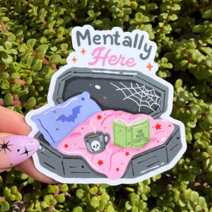 May include: A die-cut sticker featuring a dark gray coffin with a light blue pillow with a bat, a pink blanket with red stars, a gray skull mug, and a green open book with the text "ONE MORE CHAPTER" inside. The text "Mentally Here" is above.