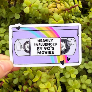 May include: A sticker featuring a purple VHS tape design with the text "HEAVILY INFLUENCED BY 90'S MOVIES" in white. The design includes a rainbow and black heart accents. The sticker is held against a background of green foliage.