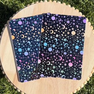 May include: Two rectangular cards featuring a black background with a repeating pattern of colorful stars, moons, and circular shapes in shades of pink, blue, purple, and yellow. The surface has a holographic finish that reflects light.