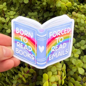 May include: A sticker in the shape of an open book with a rainbow design. The left page reads "BORN TO READ BOOKS" and the right page reads "FORCED TO READ EMAILS". A small white heart is between the pages. The sticker is held against a background of green foliage.