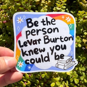 May include: A square sticker with a blue border and a rainbow arc. The sticker reads "Be the person Levar Burton knew you could be" in black text. There are small flower and star illustrations. A stack of books is in the lower right corner.
