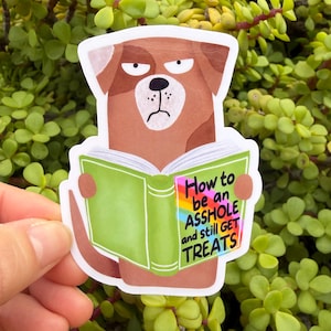May include: A sticker featuring a cartoon dog with a grumpy expression, holding a green book. The book's title reads "How to be an ASSHOLE and still GET TREATS" in colorful text. The sticker is held against a backdrop of green foliage.