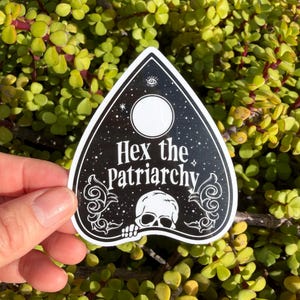 May include: A black, heart-shaped planchette sticker featuring a white skull illustration at the bottom, a large white circle at the top, and the text "Hex the Patriarchy" printed in white in the center, surrounded by decorative white filigree.