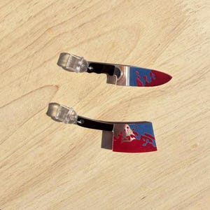 May include: Two miniature knife-shaped earrings with red and blue accents, displayed on a round wooden tray with a beaded edge. The earrings have clear plastic posts and black handles. The tray sits on a bed of green grass.