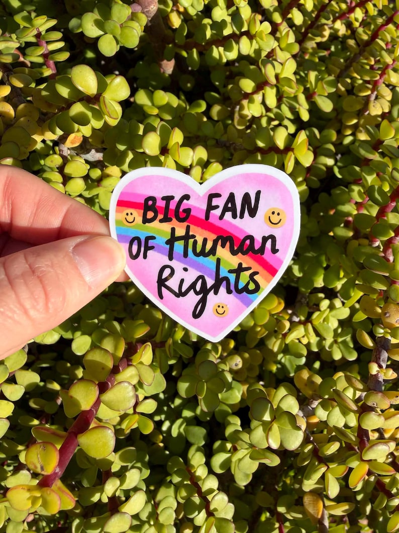 May include: A heart-shaped sticker with the text "BIG FAN OF Human Rights" in black, rainbow, and white. The sticker has a pink background, a rainbow arch, and smiling faces. The sticker is held against a background of green and yellow leaves.