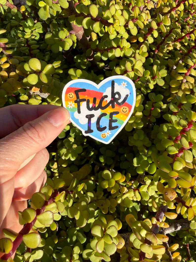 以下が含まれることがあります： 虹色のデザインと黒い文字で「Fuck ICE」と書かれたハート型のステッカー。ステッカーには笑顔と小さな花のグラフィックも付いています。ステッカーは緑の植物を背景に保持されています。