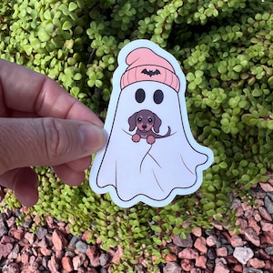May include: A die-cut sticker featuring a white ghost wearing a light pink beanie with a small black bat icon on the front. The ghost has two black oval eyes and is holding a small brown and black dachshund puppy in its center.