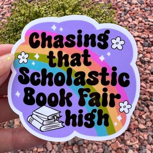 May include: A colorful sticker with the text "Chasing that Scholastic Book Fair high" in black, over a rainbow design. The sticker has a purple cloud-shaped border with white flower and diamond accents, and a stack of books.